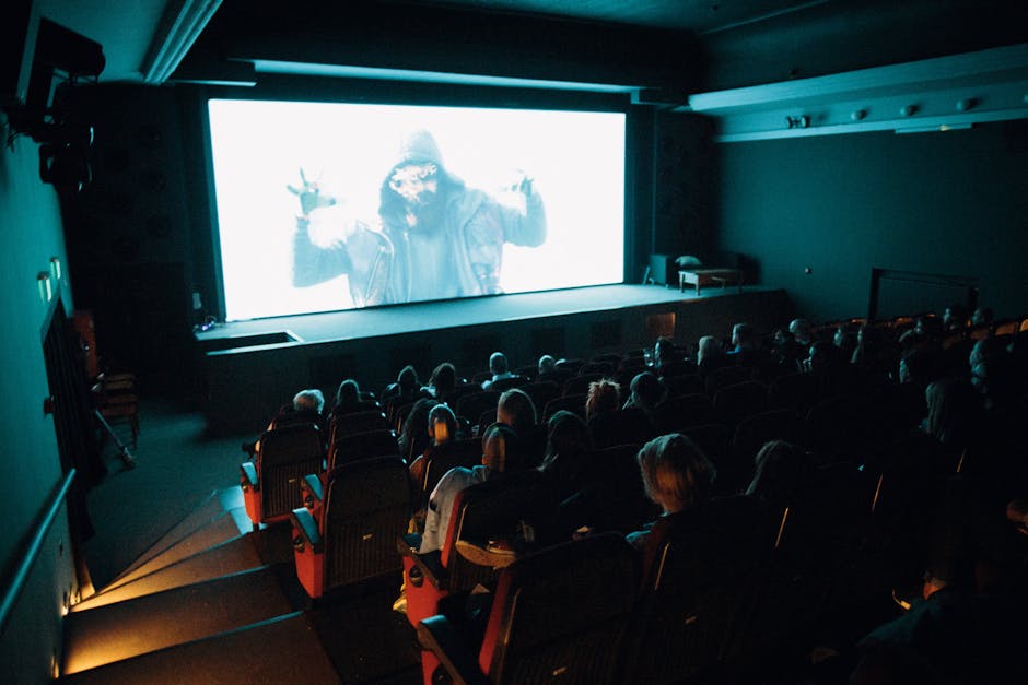 Crowded cinema theater with audience watching a movie on a large screen in a dark setting.